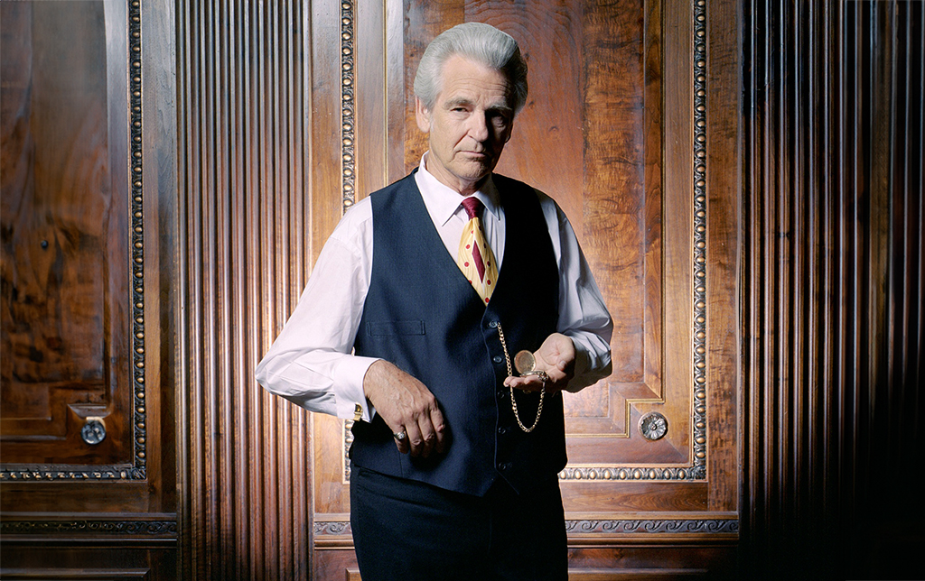 A gentleman with grey hair stands with a suit vest, tie and blue dress pants in front of a decorative wooden wall. He is holding a pocket watch.