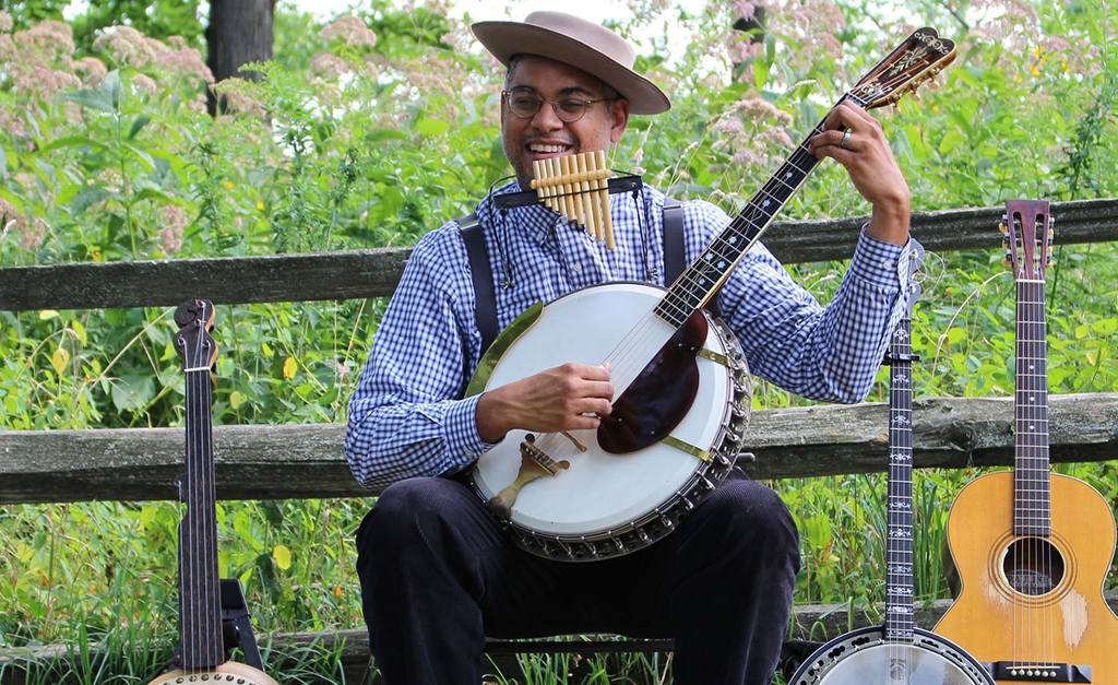 In the image, a person is sitting on a wooden bench in a park, playing a banjo. They are dressed in a blue and white checkered shirt, black pants, and a brown hat. Two guitars are resting against the bench next to them. The background consists of greenery and trees.
