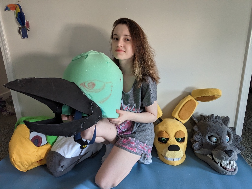 Ellie Rebellón kneels beside three created animal heads. She is wearing a gray shirt and is holding the base of the kingfisher costume head. 