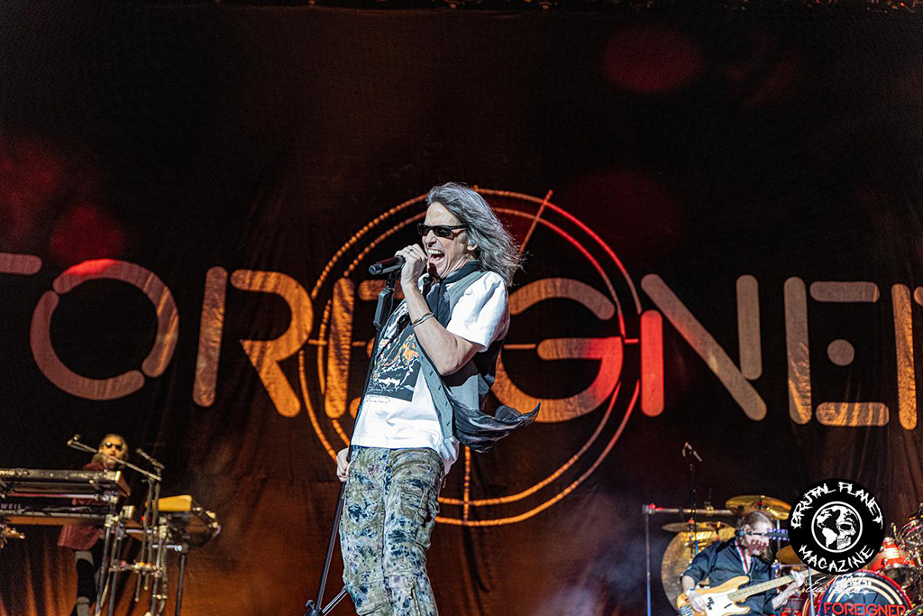 A person with long hair stands on a stage, microphone in hand. They are dressed in a white t-shirt with a black vest over it and blue jeans with a pattern. The stage is set against a large banner with the word “Foreigner” written on it, and is adorned with drums and other musical instruments.