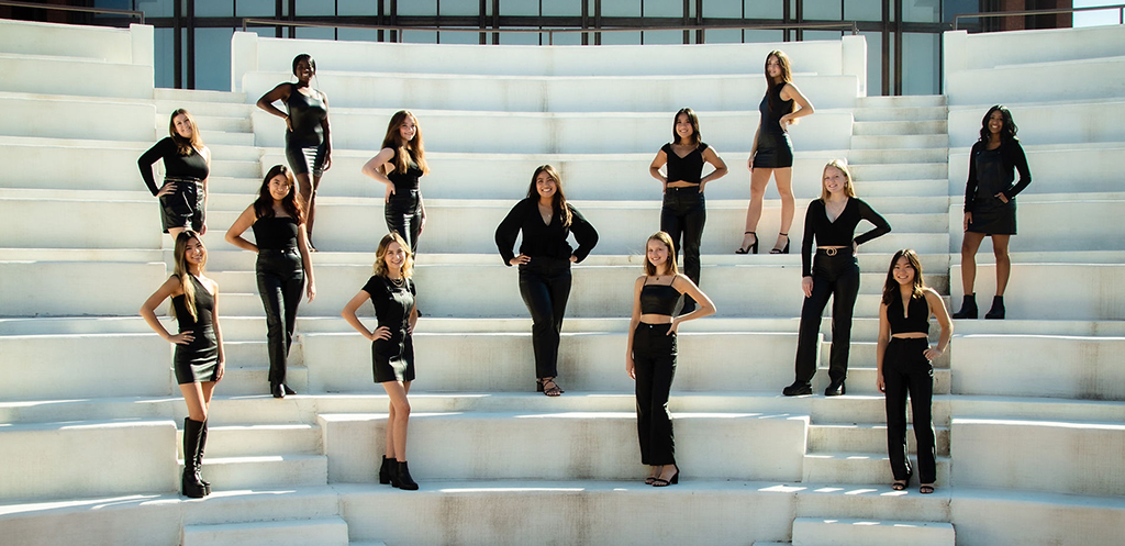 A group of individuals are assembled on a set of white stairs, each striking a unique pose. All are dressed in black attire, some standing, some sitting, and others kneeling. The backdrop is a modern building with expansive windows.
