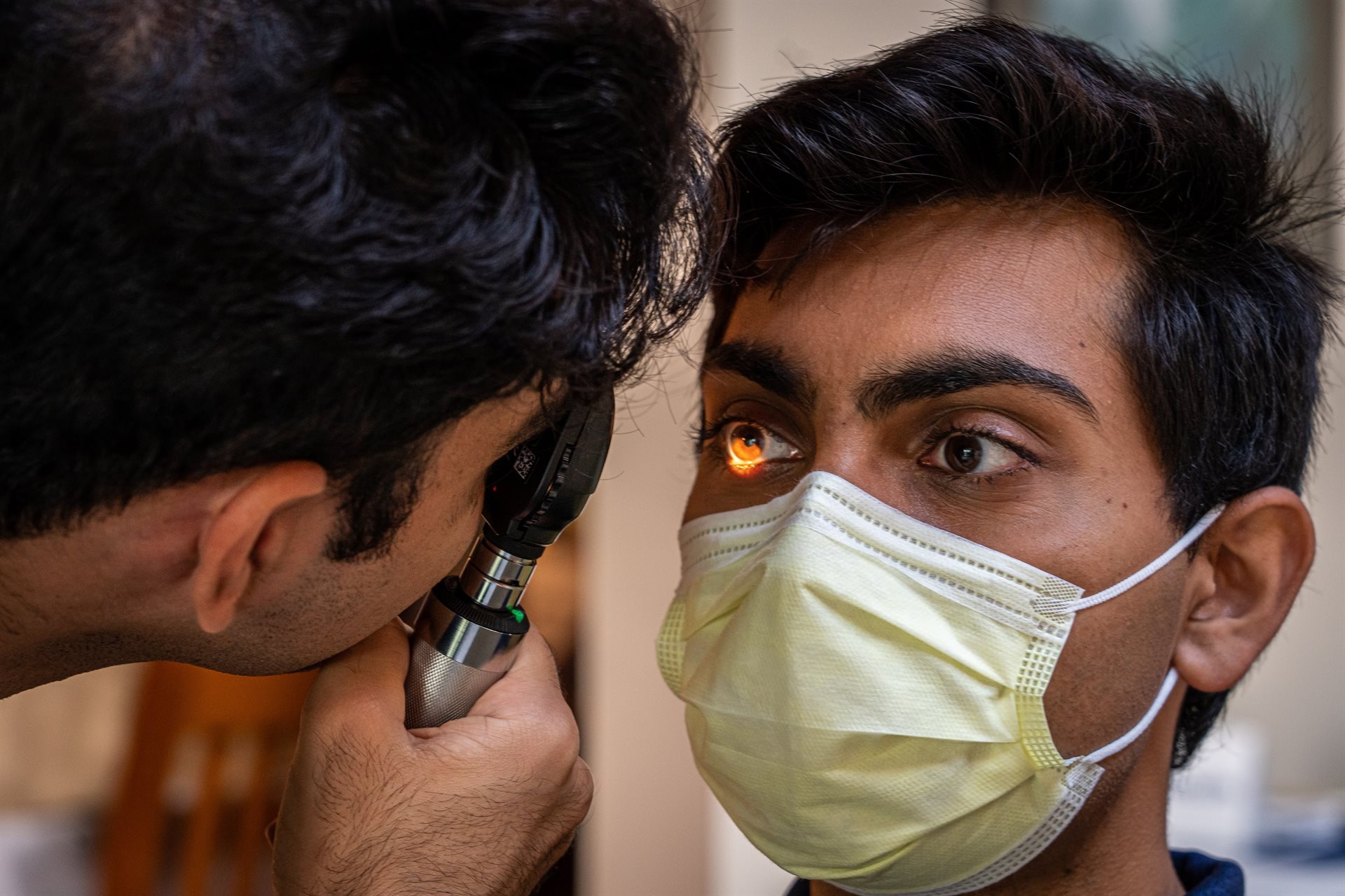A man is holding an ophthalmoscope and looking into the eye of a another man, who is wearing a surgical mask.