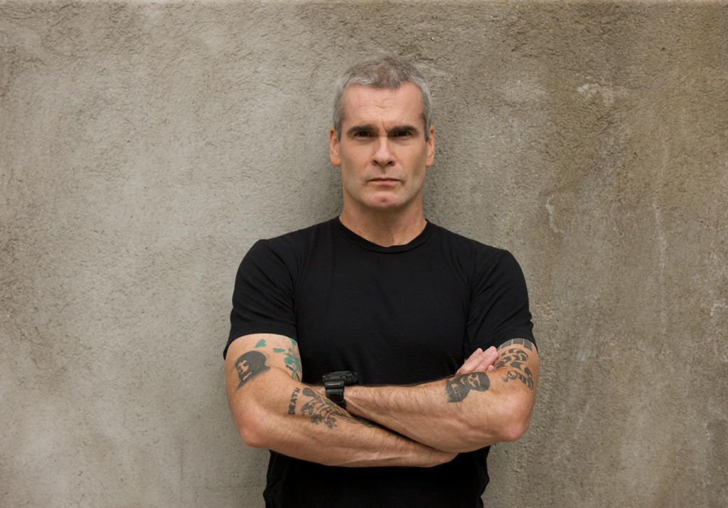 A person stands in front of a concrete wall, arms crossed. They are dressed in a black t-shirt and have tattoos on both arms. A watch adorns their left wrist.