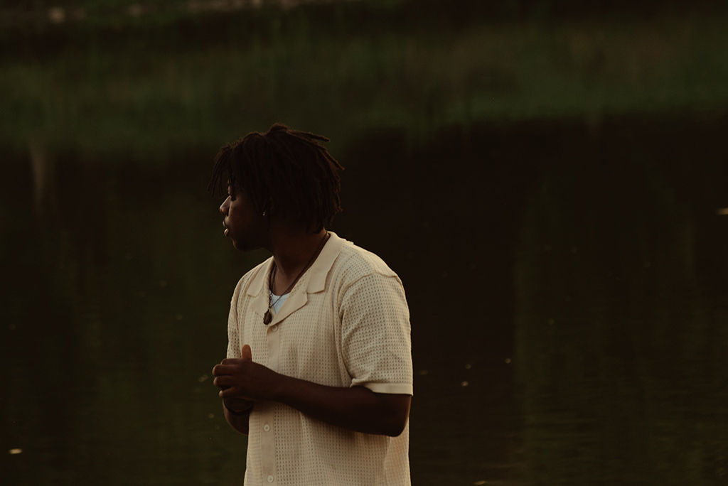 An individual is standing by a body of water. They are attired in a white shirt adorned with a necklace. Their hair is styled in dreadlocks. Their hands are clasped together in front of them. The backdrop consists of a serene body of water and lush greenery.