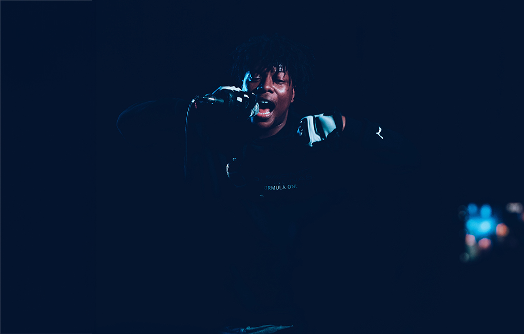 A Black man is holding a microphone and appears to be rapping into it in a dark room. They are dressed in a black jacket. In the distance, colorful lights can be seen.