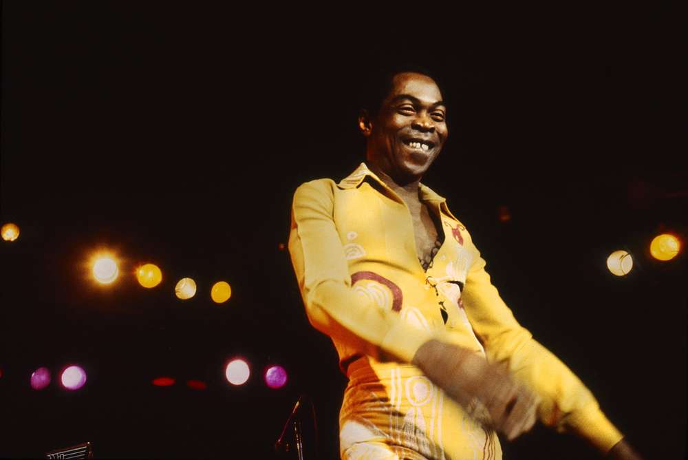A smiling Black man in a yellow jumpsuit, standing on a stage with lights behind him.