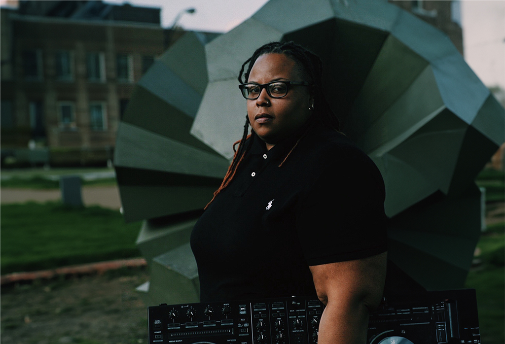 A person is standing in front of a large, geometric sculpture. They are wearing a black polo shirt with a white logo on the left chest and holding a large, black DJ mixer. The sculpture consists of multiple triangular and rectangular panels in varying shades of gray. It’s situated on a grassy lawn with a brick building in the background.