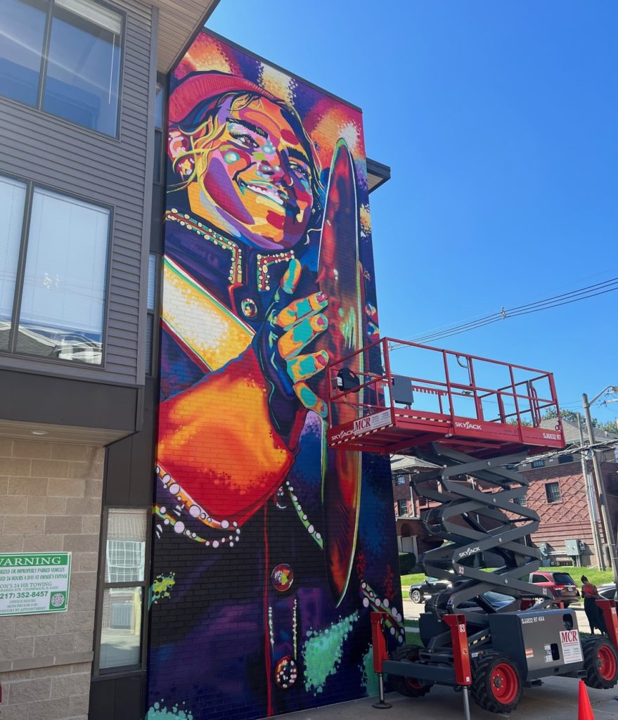 A brightly colored mural depicting a cymbal player in a marching band uniform. It stretches from the top to the bottom of the wall of an apartment building. There is scaffolding alongside it.