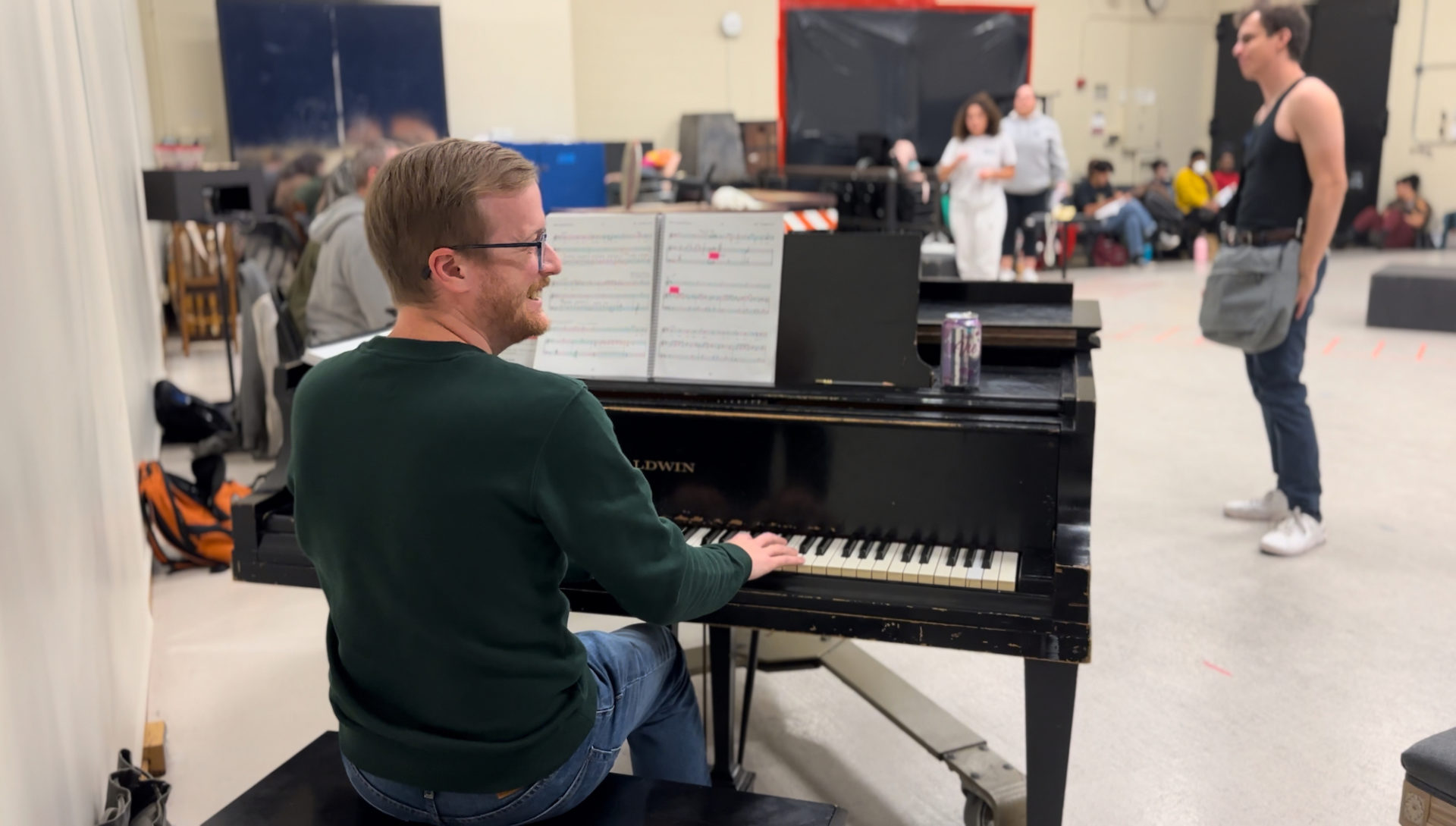 Justin Brauer, a white man with light hair and glasses, sits at a piano in a rehearsal space