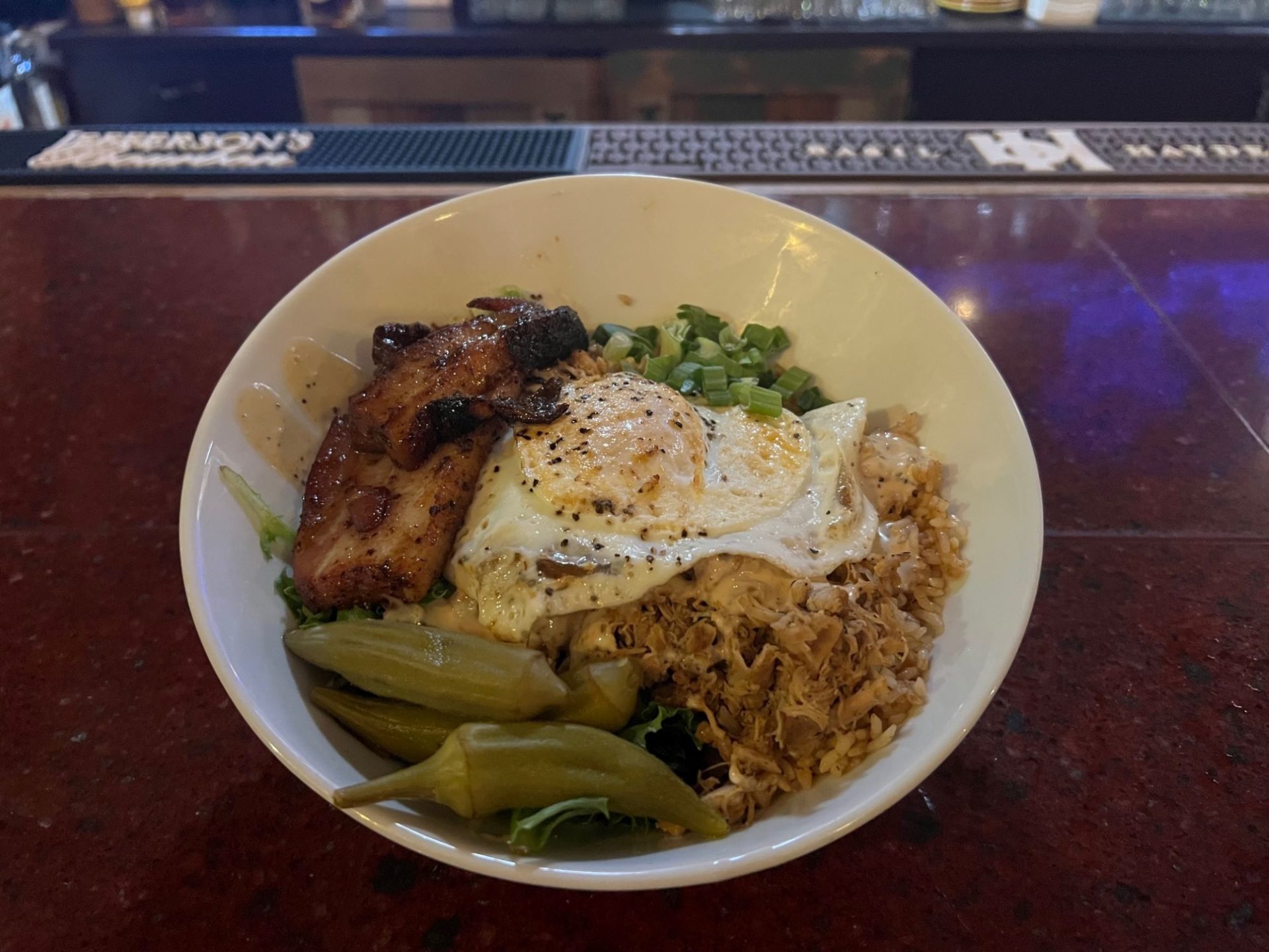 A white bowl filled with rice, pulled chicken, pork belly, green onions, pickled okra, pork belly, and a fried egg.