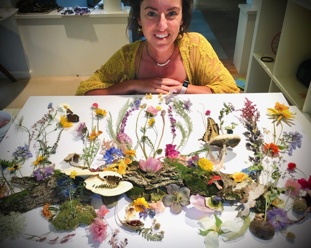 Cris Hughes, a young white woman, sits behind a table smiling with an arrangement of dried flowers laid out on a white table in front of her 