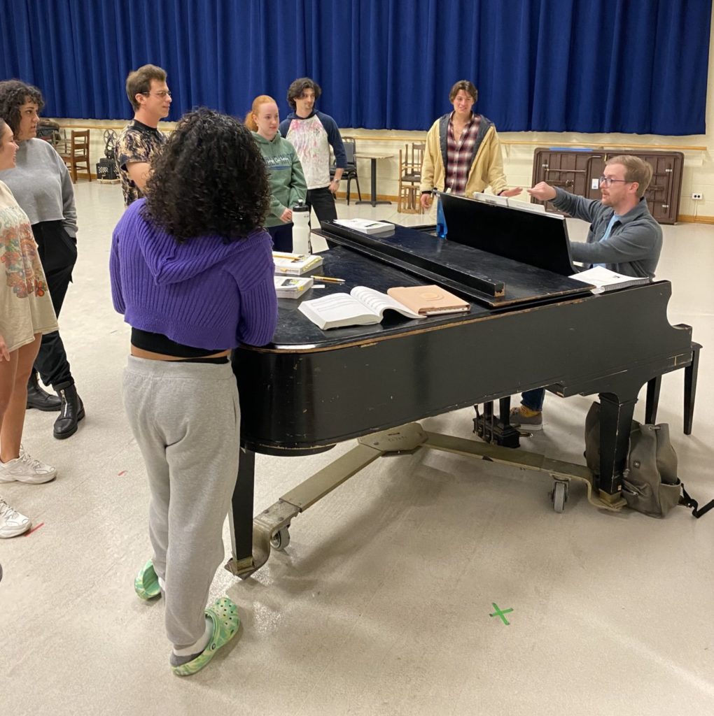 Justin Brauer, a white man with light hair and glasses, sits at a piano in a rehearsal space with actors singing around him
