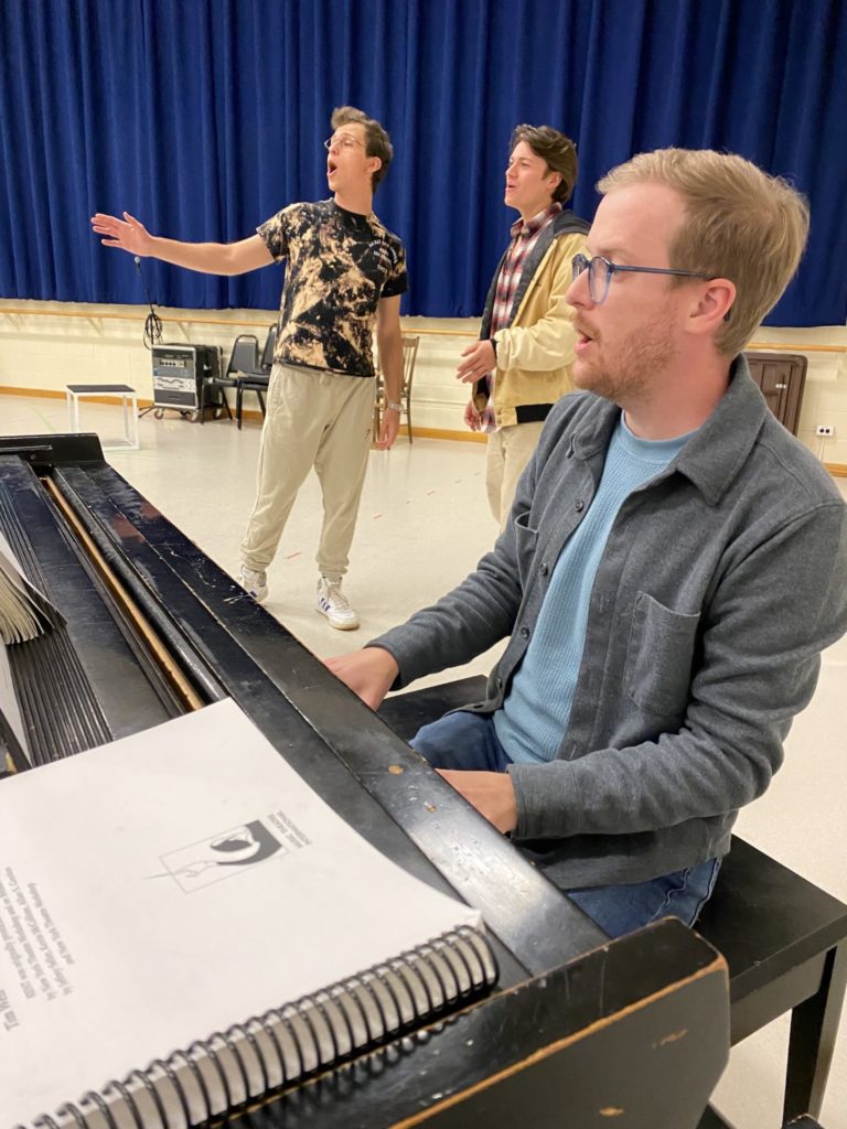 Justin Brauer, a white man with light hair and glasses, sits at a piano in a rehearsal space with actors visible singing behind him