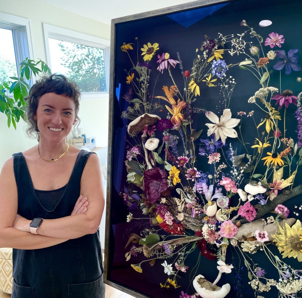 Cris Hughes, a white woman, stands next to the large completed community art project: a black large square box filled with colorful dried flowers