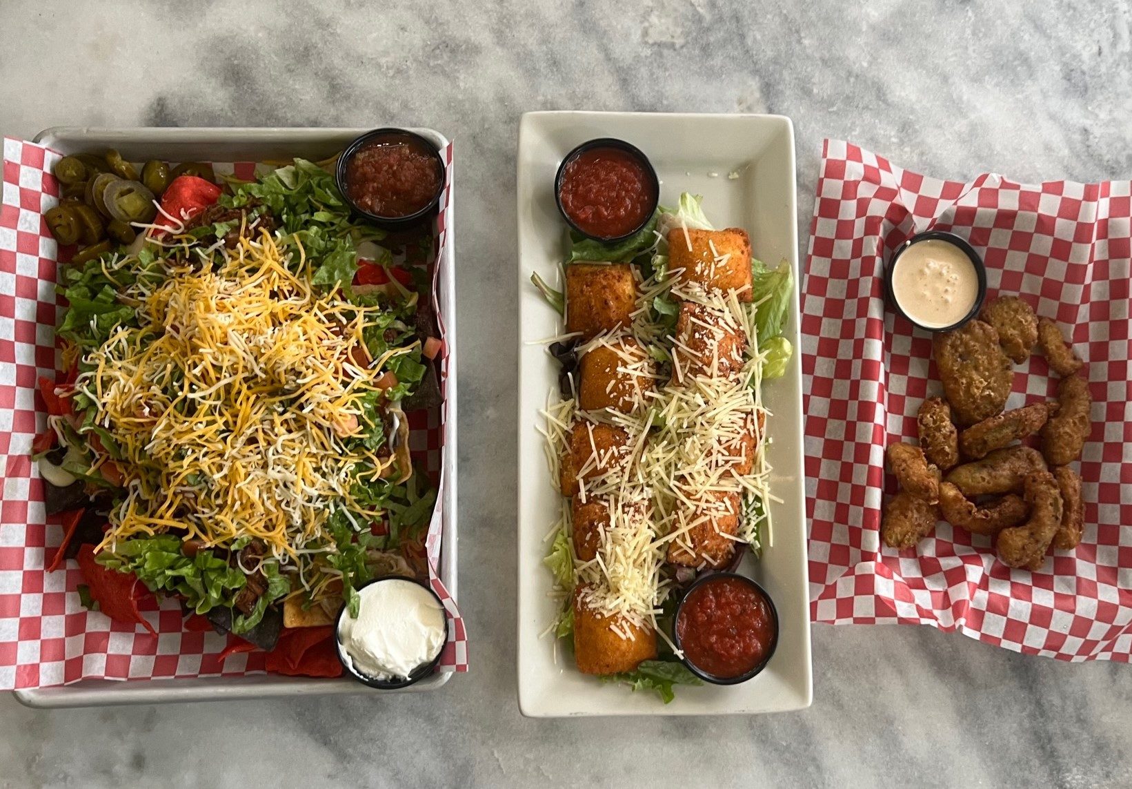 A row of three appetizers: nachos, toasted ravioli, and deep fried avocado slices.