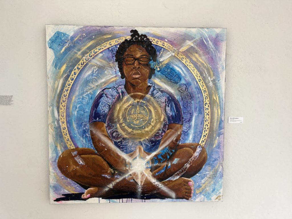 square painting of a Black person sitting legs crossed with hands in front and eyes closed. They are surrounded by a gold circle, and there is a light coming from where their hands are joined. 