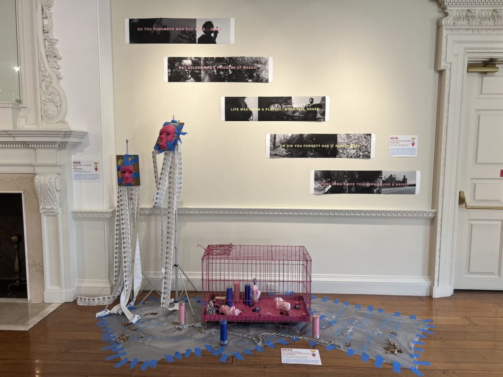 a hot pink dog cage sits on the floor with statues of the virgin mary scattered about inside along with religious candles. There is a chain on the floor, two plaster face masks on stands, and quotes with photographs printed and hanging in strips on the wall. 