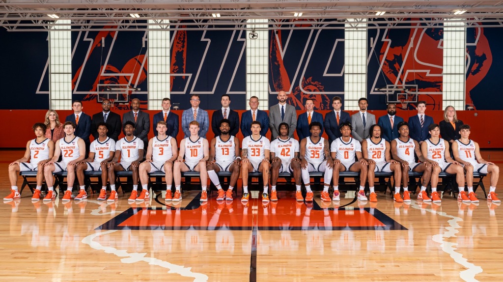 The men's basketball team sits in chairs in a straight line on the basketball court. They are wearing white jersey's with orange lettering. Behind them in a second row, is a row of coaches wearing suits and ties. They are all men, except for two women on each side of the end of the back row.