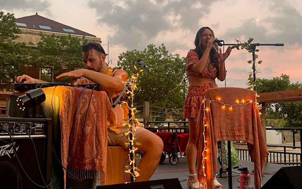 Two people are on a stage with musical instruments. The person on the left is sitting on a stool and playing a guitar, wearing a yellow shirt and shorts. The person on the right is standing and singing into a microphone, wearing a red dress with a floral pattern. The stage is decorated with lights and plants. The background consists of a building and trees. The image is taken during the day.