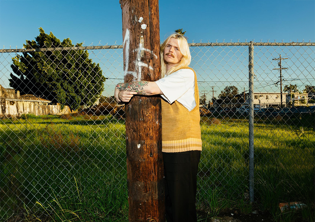 A person with long blood hair and a mustache hugs a telephone pole in front of a chain link fences in front of an empty lot in a gritty neighborhood. The subject is wearing a white t-shirt with a yellow sweater vest and black pants.