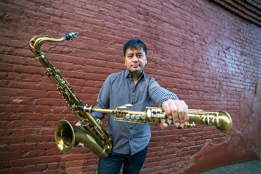 An individual is facing forward, their striped shirt and the golden saxophone they hold forming a striking contrast against the backdrop of the weathered brick wall. The anticipation of a melody fills the air, even in the stillness of the image.