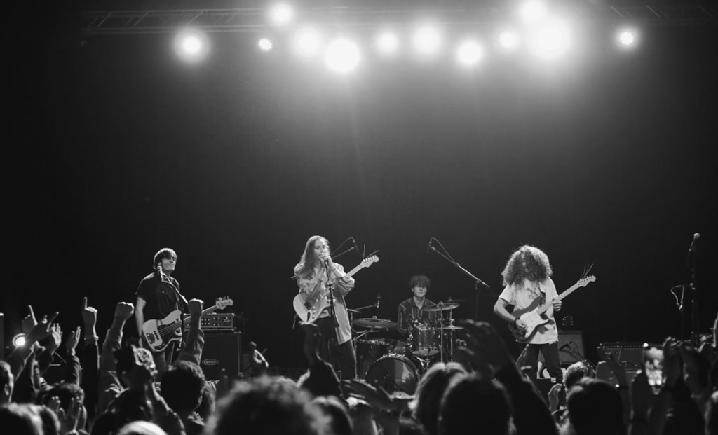 A black and white photo of a rock band on stage. Above you can see the stage lights shining bright, and below you see the audience enthusiastically raising their fists and singing.