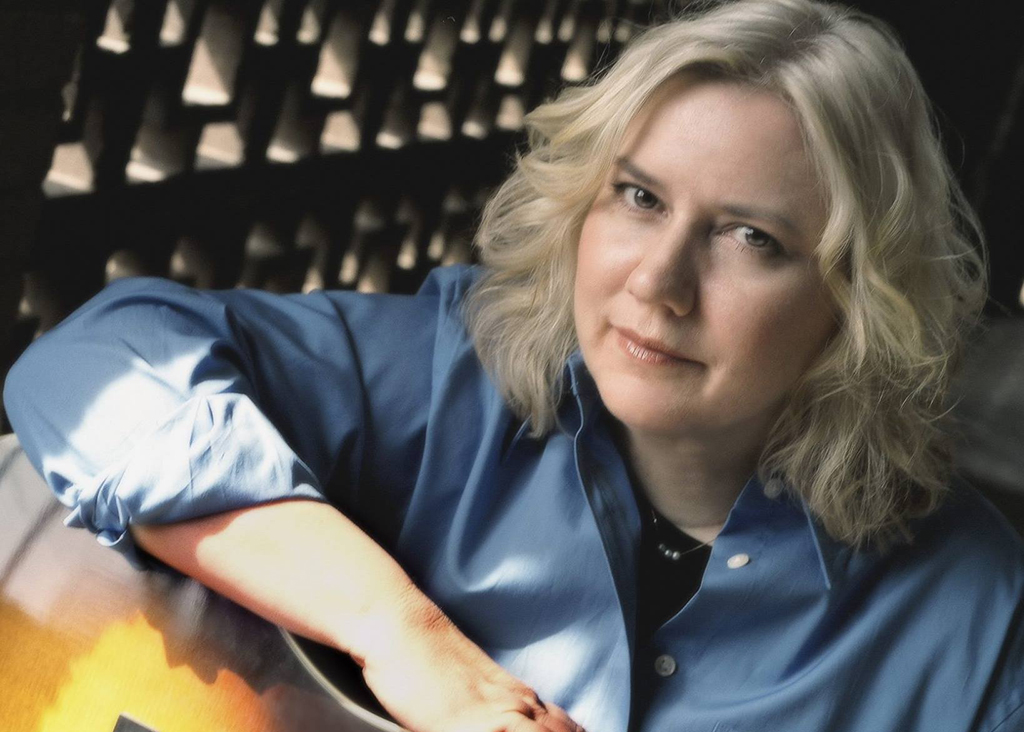 A person is holding a guitar, attired in a blue button-down shirt with the sleeves rolled up. Their hair is blonde and falls to shoulder-length.