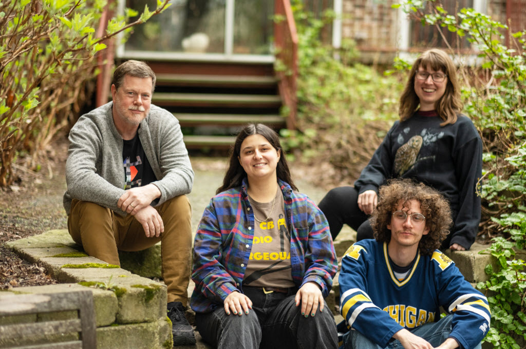 Four individuals are captured in a relaxed and casual setting. They are seated on a stone staircase surrounded by a garden. They are dressed in casual attire, including sweatshirts and t-shirts. The background features a wooden fence and an array of plants. The overall mood conveyed is one of leisure and ease.