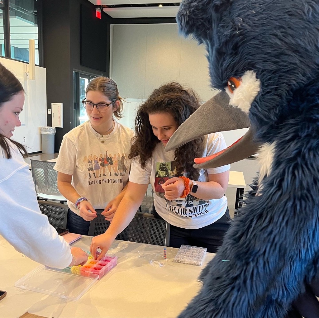 Three girls in Taylor swift t-shirts stand around the table with the Kingfisher mascot. They appear to be making friendship bracelets.