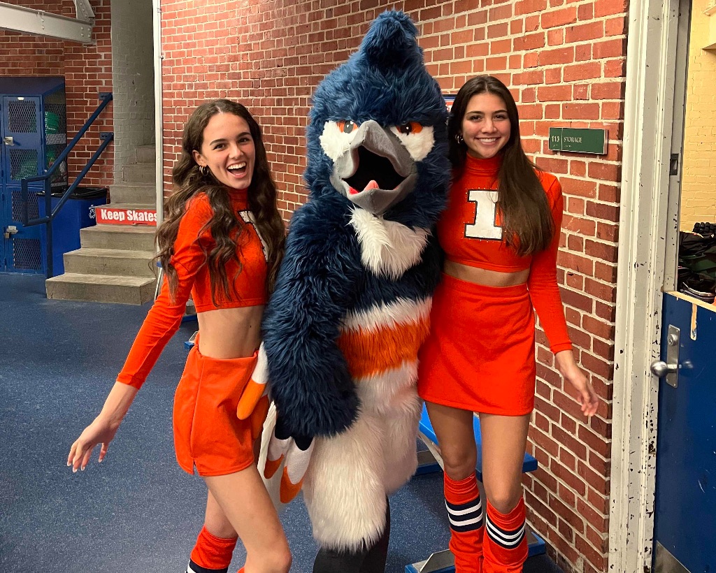 Two woman with long brown hair and matching orange cropped shirts with white I'd and orange skirts and socks stand next to the Kingfisher mascot in front of a brick wall. 