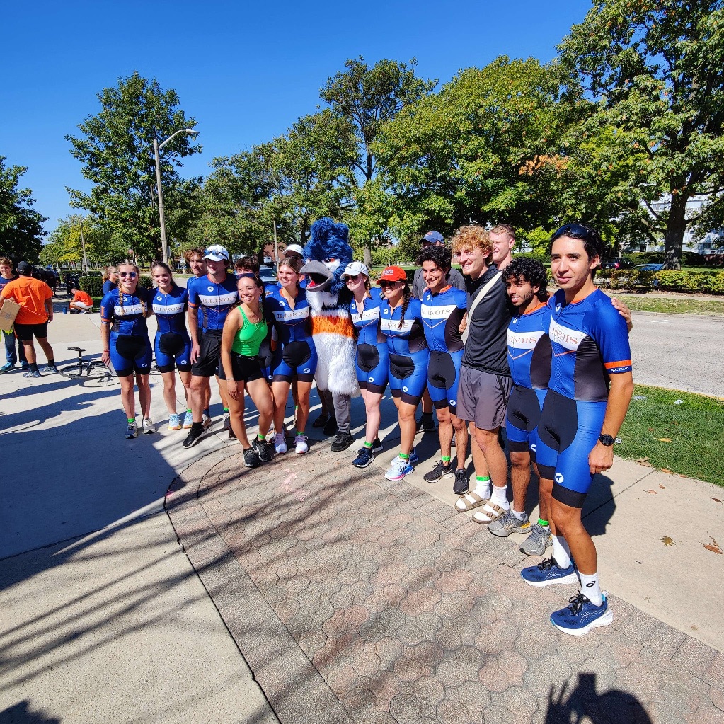 A group of students stand in a line on a sidewalk smiling at the camera. They are all wearing matching blue cyclingoutfits and the Kingfisher mascot standing with them.