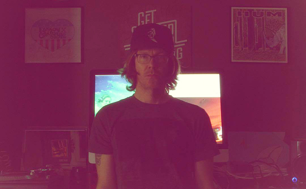 A person is standing in front of a desk that supports two computer monitors. They are attired in a black t-shirt and a baseball cap. The desk is populated with a keyboard, a mouse, and a few other items. The wall behind the desk is adorned with posters and a clock.