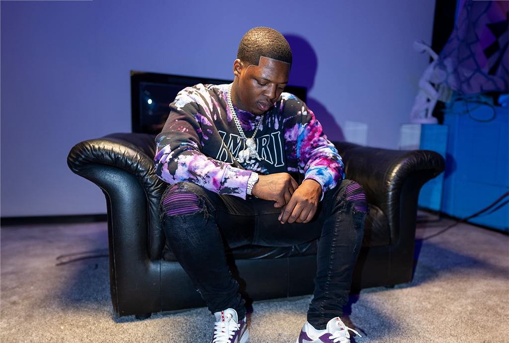 A person is sitting on a black leather couch in a room with a purple wall and a TV in the background. They are wearing a multicolored hoodie with the word “MIRI” on it, ripped jeans, and white sneakers. Their legs are crossed and their arms are resting on their knees. The background consists of a TV, a white table, and a purple wall with a mural on it.