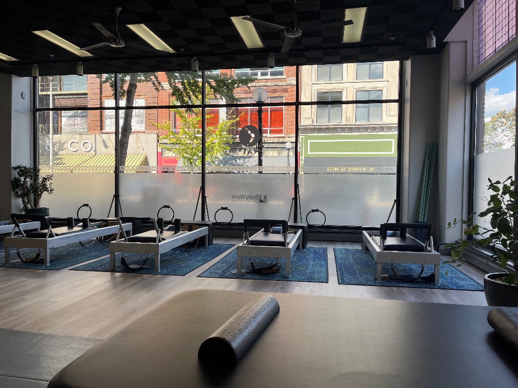 A pilates studio with pilates reformer machines on individual rugs in front of half frosted tall windows. There are buildings visible through the window. 