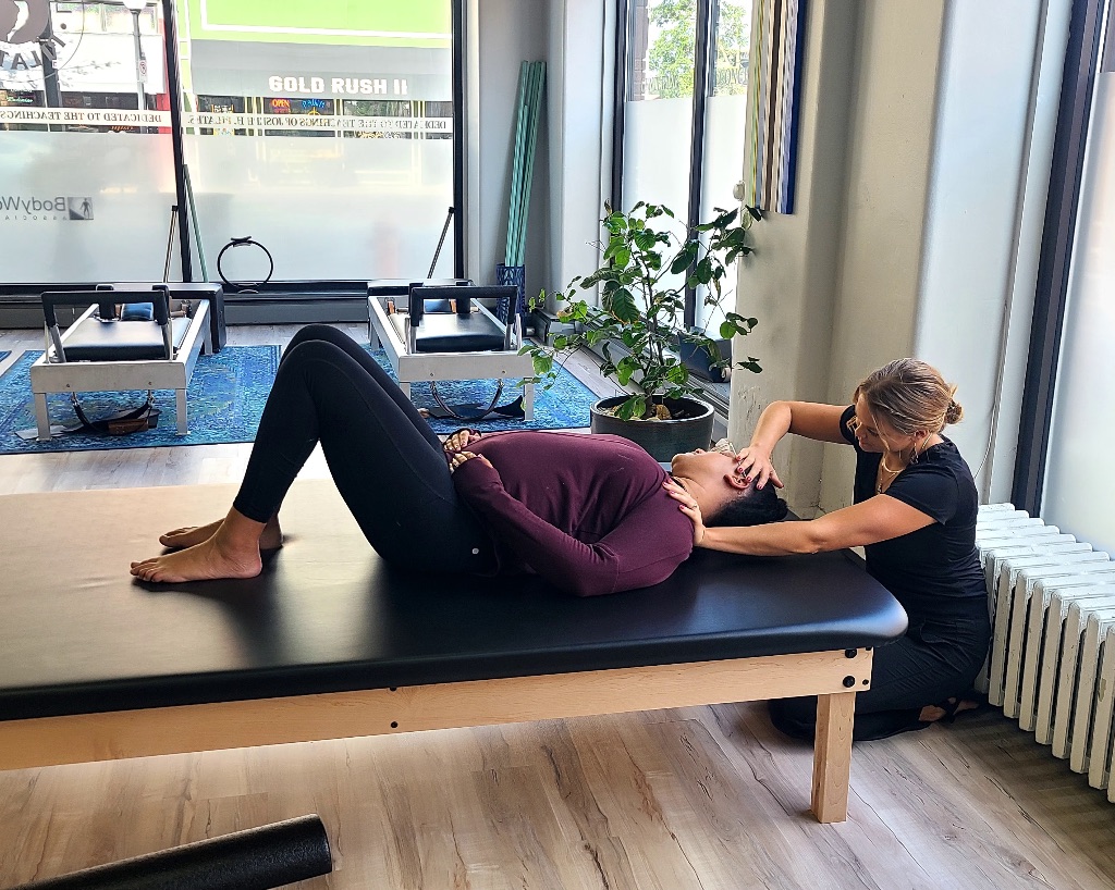 In a large pilates studio a blond woman in a black t-shirt holds the shoulders of a woman laying on a padded black table with wooden legs.