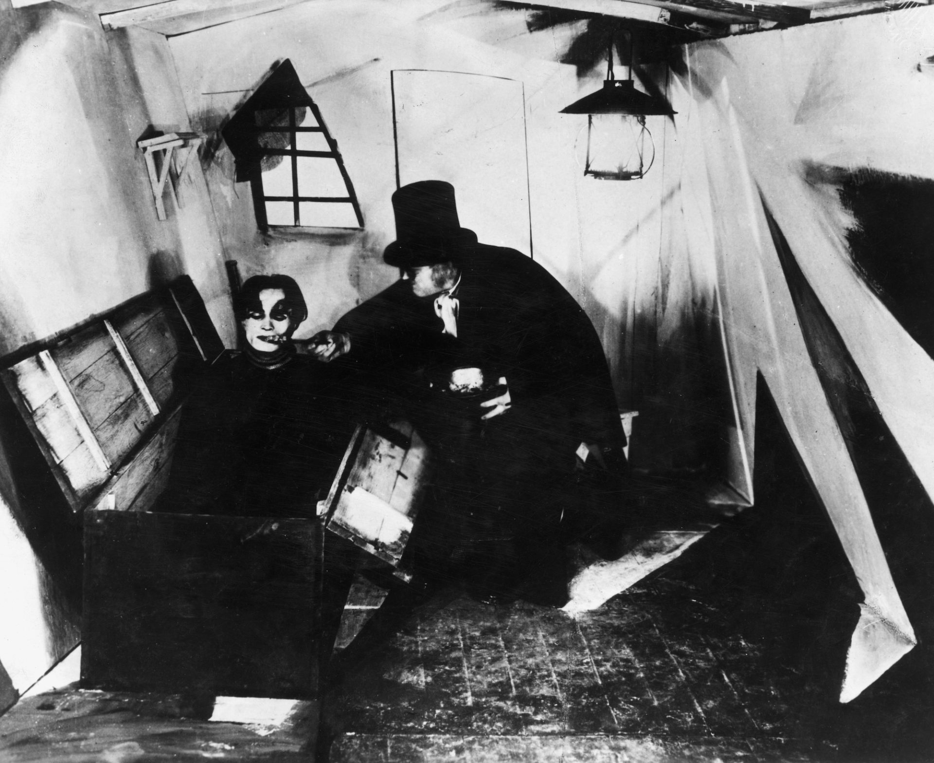 A scene from The Cabinet Of Dr Caligari in black and white shows a man in a top hat and black cape leaning over another man emerging from a casket