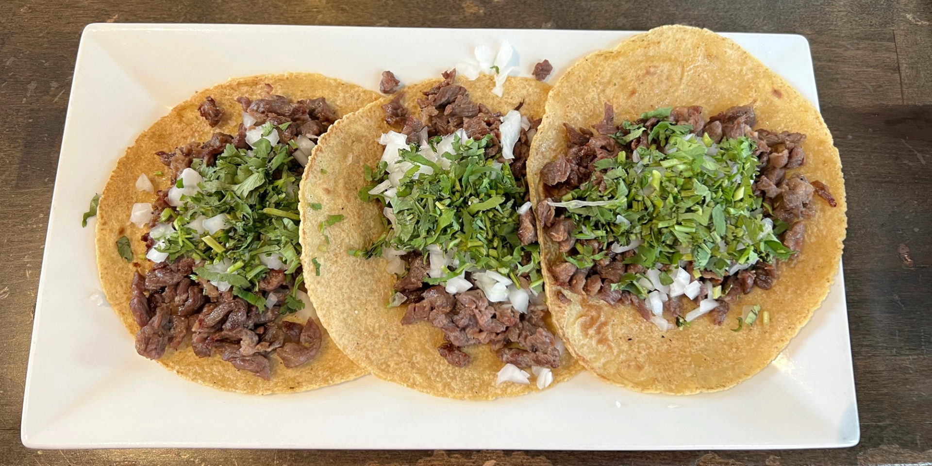 Three carne asada tacos with cilantro and onions on soft corn tortillas on a white rectangular plate at Maize Mexican Grill in Champaign, Illinois.