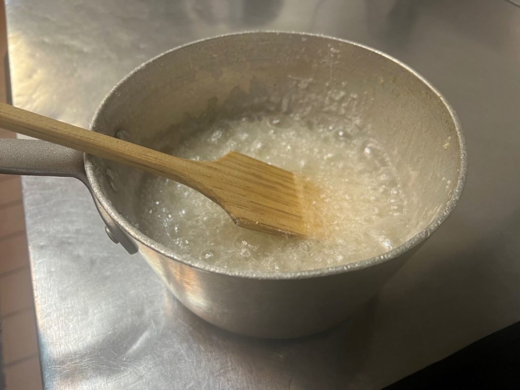 A mixture of sugar and water is bubbling in a metal pot, as it is stirred with a flat wooden utensil.