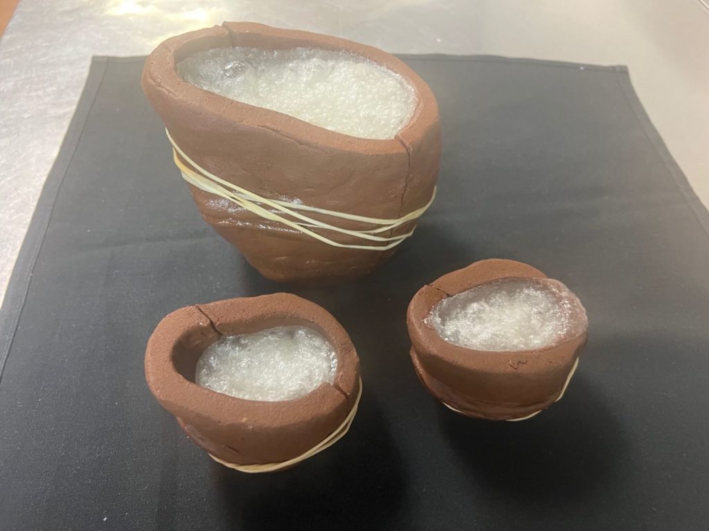 One large and two small clay molds are sitting on a black cloth. They are filled with a bubbling mixture of sugar and water and wrapped with rubber bands.