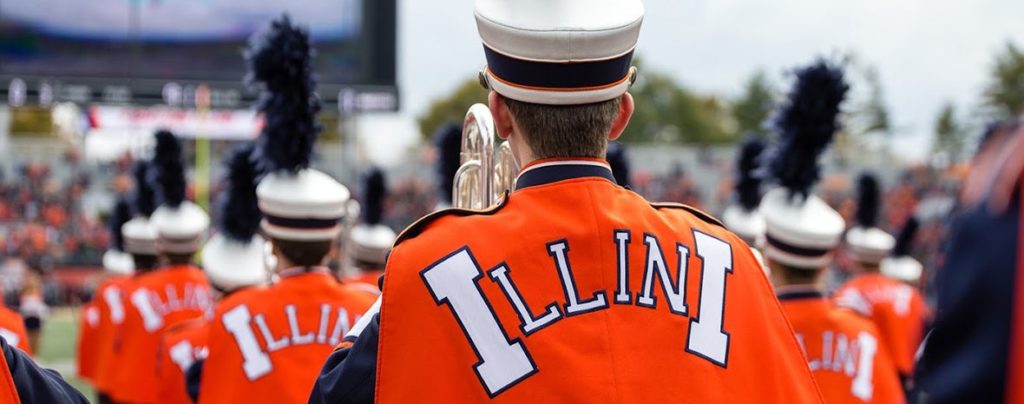 The image you uploaded appears to be of a marching band. The band members are wearing orange and white uniforms with “Illini” written on the back. They are also wearing white hats with black plumes. The photo seems to have been taken in a stadium filled with spectators, and it’s taken from behind the group, so only the backs of their heads and uniforms are visible. It’s a very dynamic and lively scene