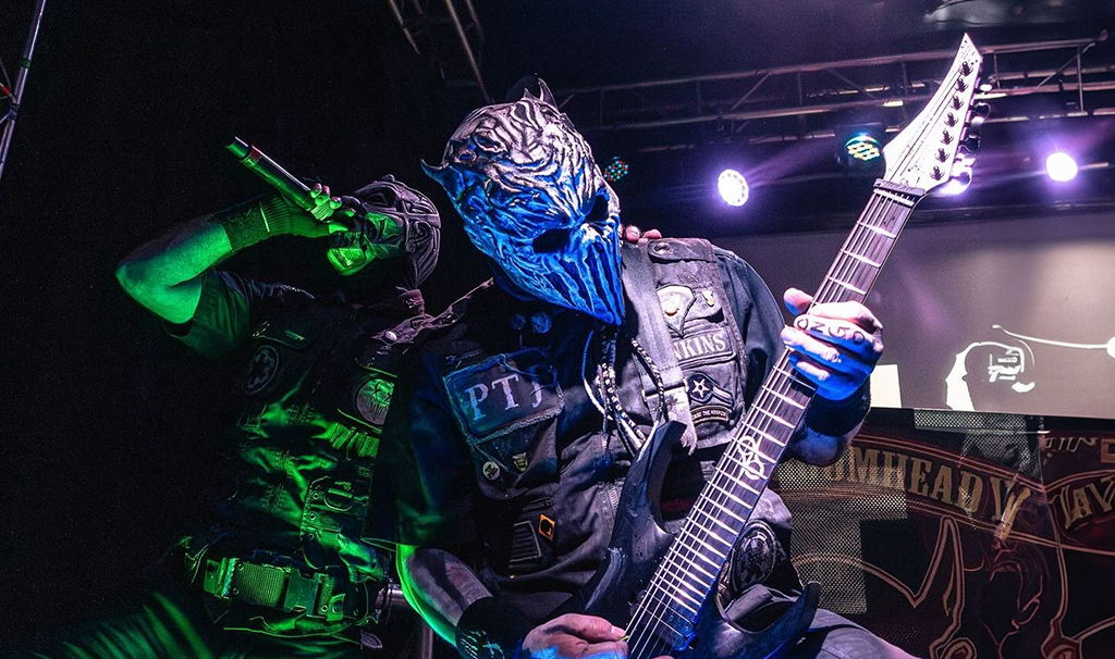Two individuals are on a stage. One is strumming a guitar, and the other is holding a microphone. They are both donning masks: one mask resembles a blue alien, and the other appears to be a green gas mask. Their attire is black, adorned with various patches and pins. The stage behind them is dimly lit with hues of purple and blue.
