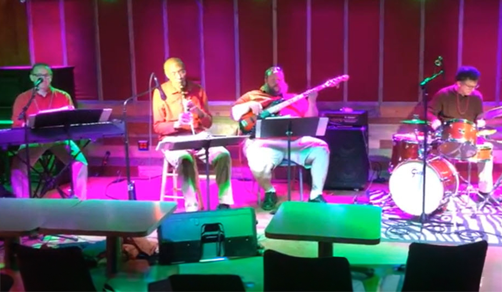 A band is performing on a stage. The stage is lit with purple and pink lights. There are four people in the band, one playing the keyboard, one playing the guitar, one playing the drums, and one playing the bass. They are all dressed in casual clothes and are sitting on stools or chairs. The stage has a wooden floor and there are empty chairs in the foreground.