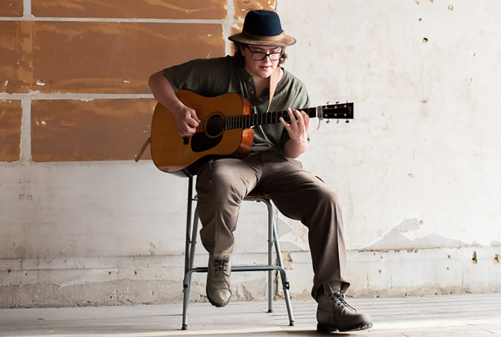 A person is sitting on a stool, playing an acoustic guitar. They are dressed in a green hat, a beige shirt, and beige pants. They are also wearing boots. The guitar is a light brown color with a black neck. The wall behind them is made of bricks and is partially painted white. The floor is made of concrete.