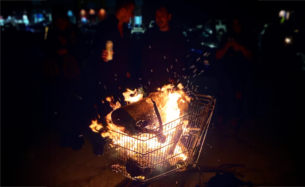 The scene is set in a dark street. A shopping cart, filled with burning trash and debris, is the focal point. The fire is bright, casting light on the surrounding area. In the background, there are indistinct figures of people and cars, adding to the overall dark and chaotic mood of the image.