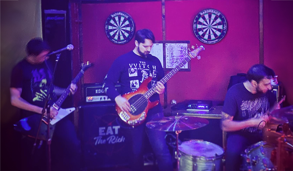 The scene unfolds on a stage in a bar. A band is performing, their energy palpable even in the still image. One member is engrossed in playing a guitar, another is behind a drum set, and a third is wielding a bass. The bass player’s black t-shirt stands out, emblazoned with the words “The Witching Hour”. The drum kit sits in front of a black banner that boldly proclaims “Eat The Rich”. The image is vibrant with the raw energy of live music.