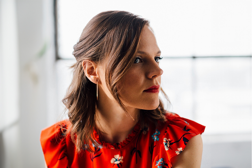 In the image, a person is seated in a room with a window casting soft light. They are dressed in a red floral dress that adds a pop of color to the scene. Their shoulder-length brown hair frames their silhouette as they face towards the right side of the image, creating an atmosphere of quiet contemplation.