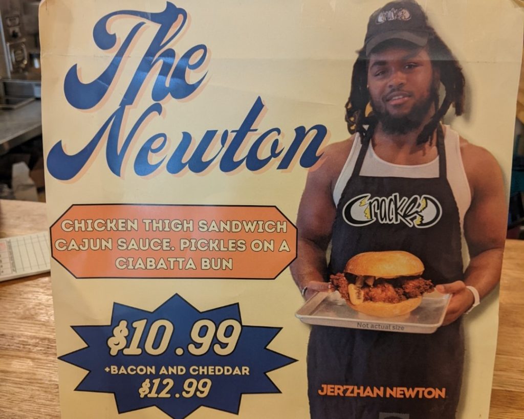 A sign that reads "The Newton, chicken thigh sandwich cajun sauce, pickles on a ciabatta bun, $10.99" with a photo of University of Illinois football player Jer'Zhan Newton wearing a cracked hat and apron and holding a tray with the sandwich.