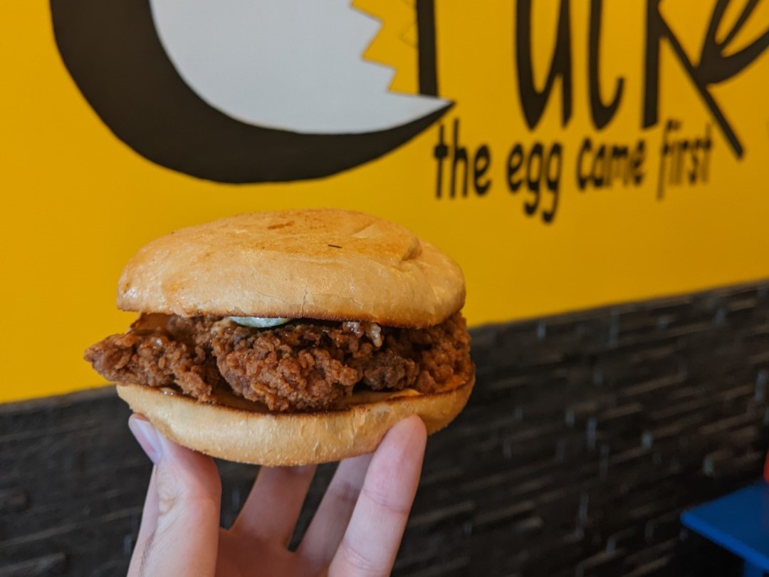 A fried chicken sandwich held by a white person's hand in front of Cracked's yellow sign inside the restaurant.