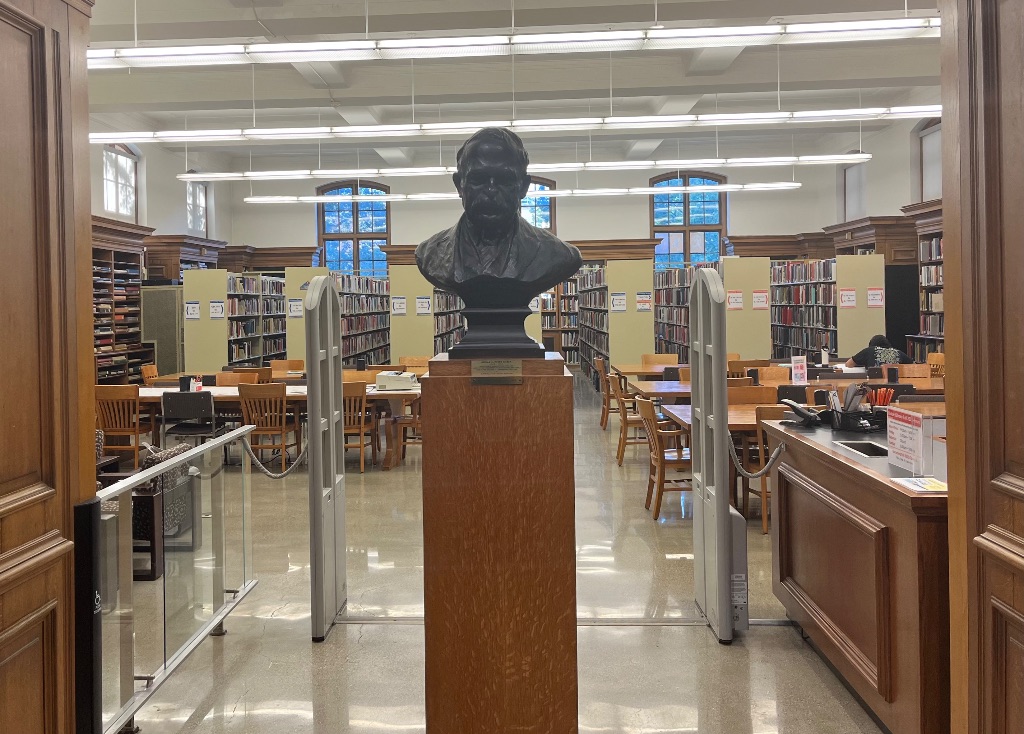 The entrance to the Ricker Library. There is a black metal bust of a man with a mustache and behind him are shelves of books and three big windows.