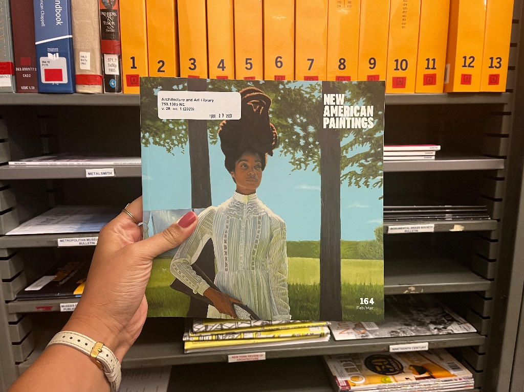 A brown hand holding a magazine in front of a metal bookshelf full of magazines. The magazine is called New American Paintings and has a painting of a black woman in a long sleeves white dress holding a gun. She has long coils of hair wrapped in a bun and she is standing in front of two drees on a sunny day.